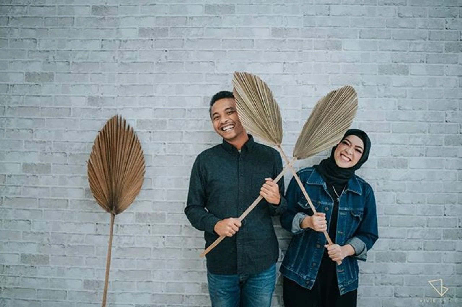Pasangan prewedding berpakaian denim memegang daun palem kering di depan dinding bata putih