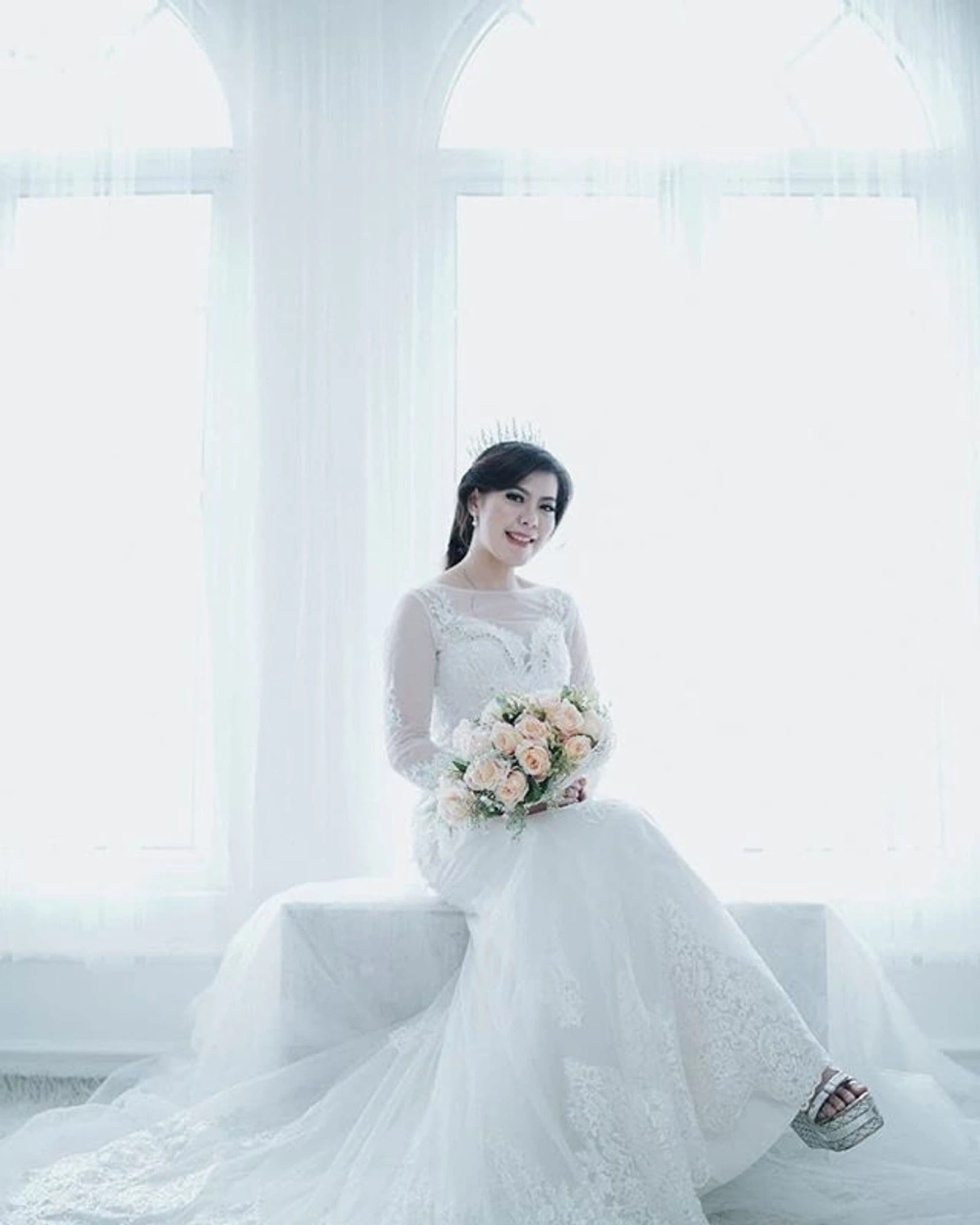 Calon pengantin wanita duduk di dekat jendela memakai gaun putih