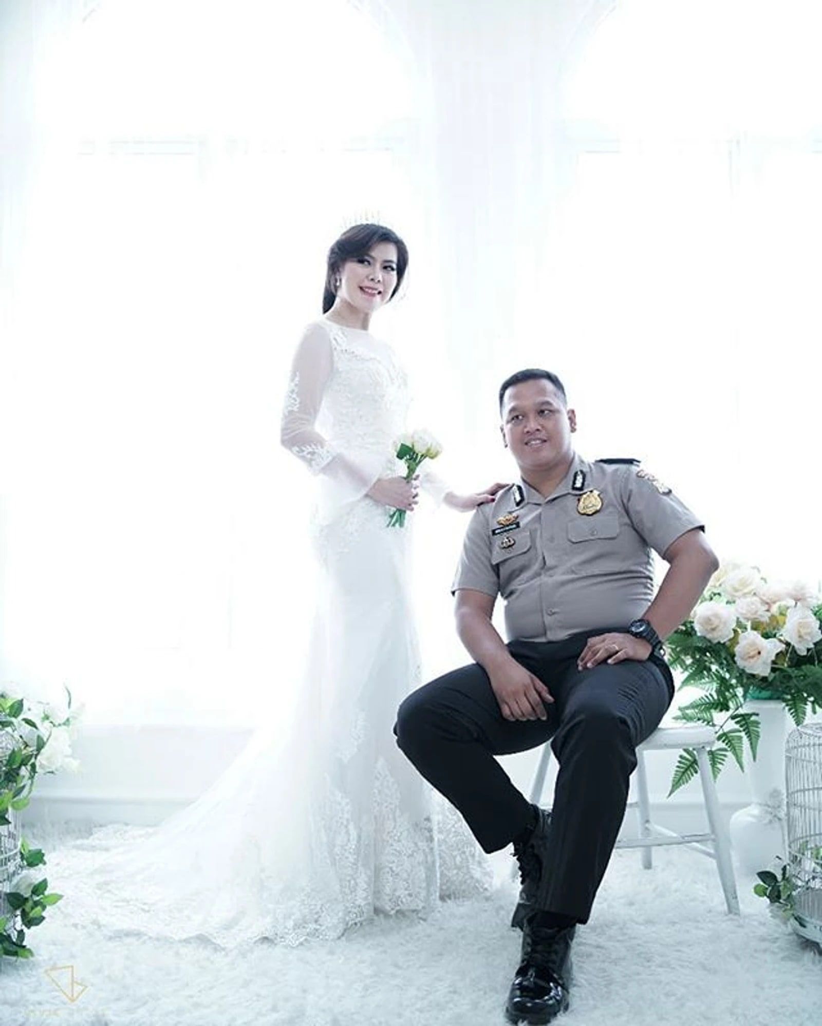 Pasangan prewedding di studio, wanita berdiri memakai gaun putih di samping pria yang duduk