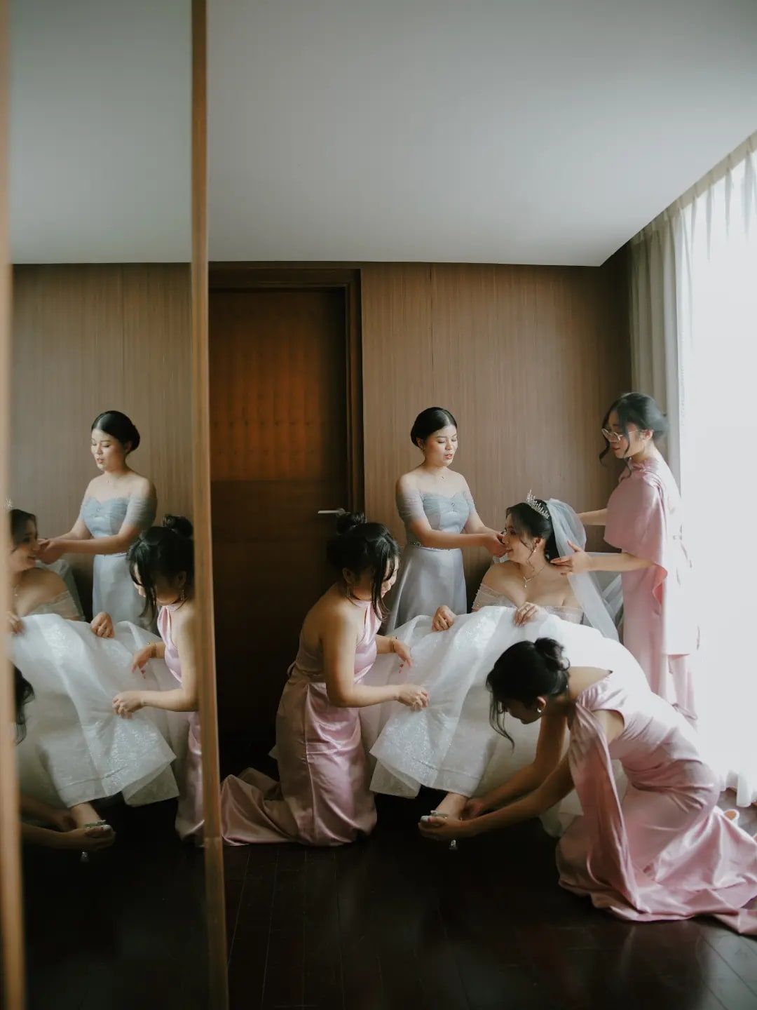 Para bridesmaid bergaun pink merapikan gaun pengantin, terlihat dari pantulan cermin
